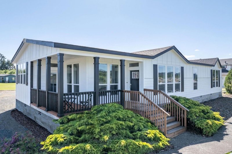 A white manufactured home with charcoal trim and a covered front porch, a wooden entry ramp rising to the door, and low juniper shrubs planted across the front